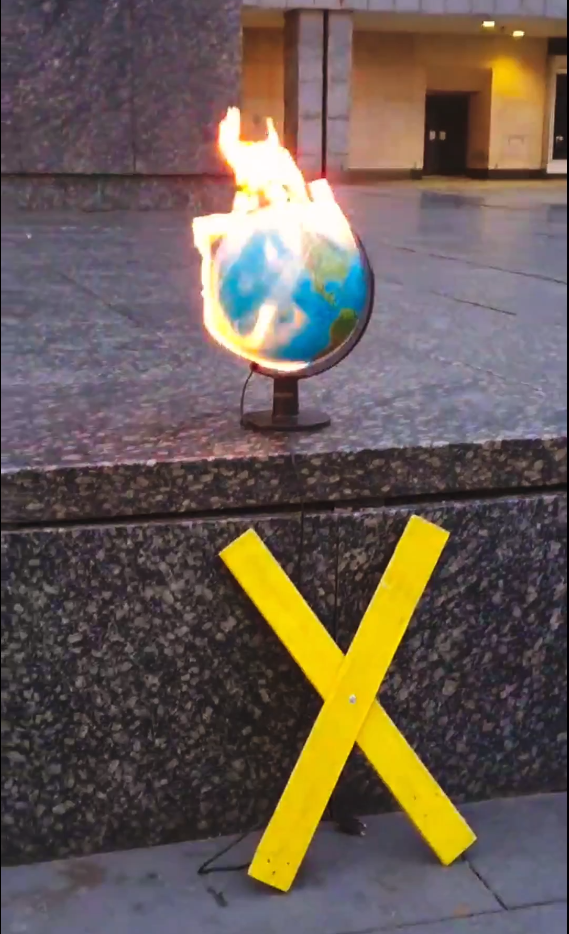 Brennender Globus vor dem Karl-Marx-Kopf. Davor ein gelbes X.